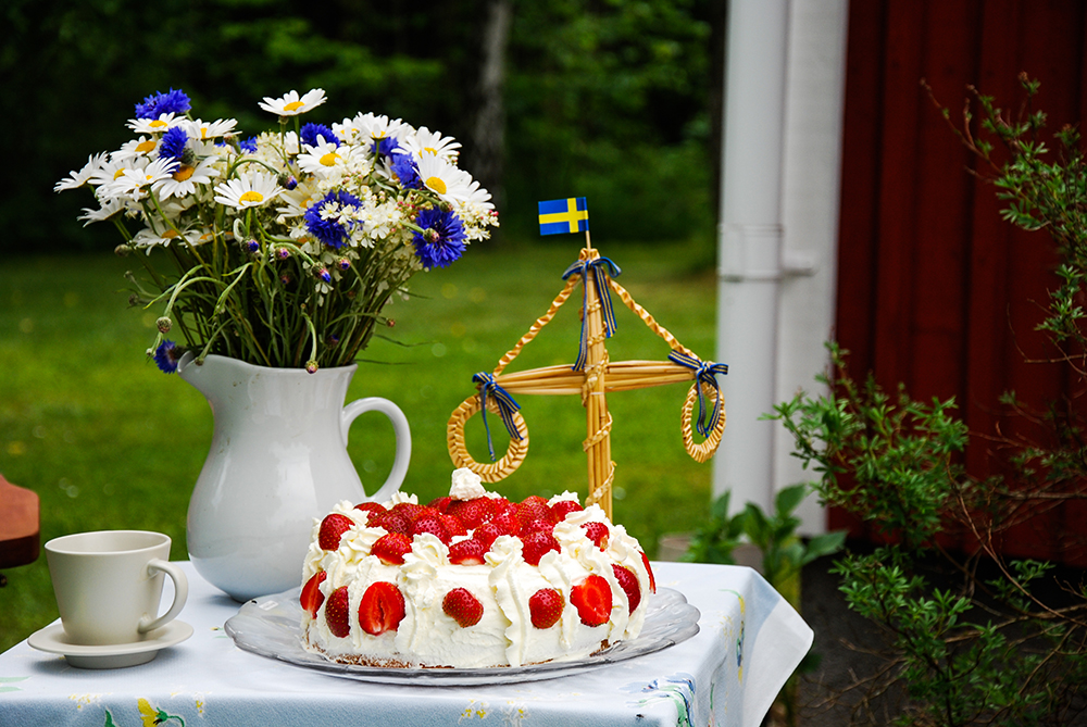 Ett bord med en sommarbukett, liten midsommarstång och en jordgubbstårta, på ett bord i en somrig t