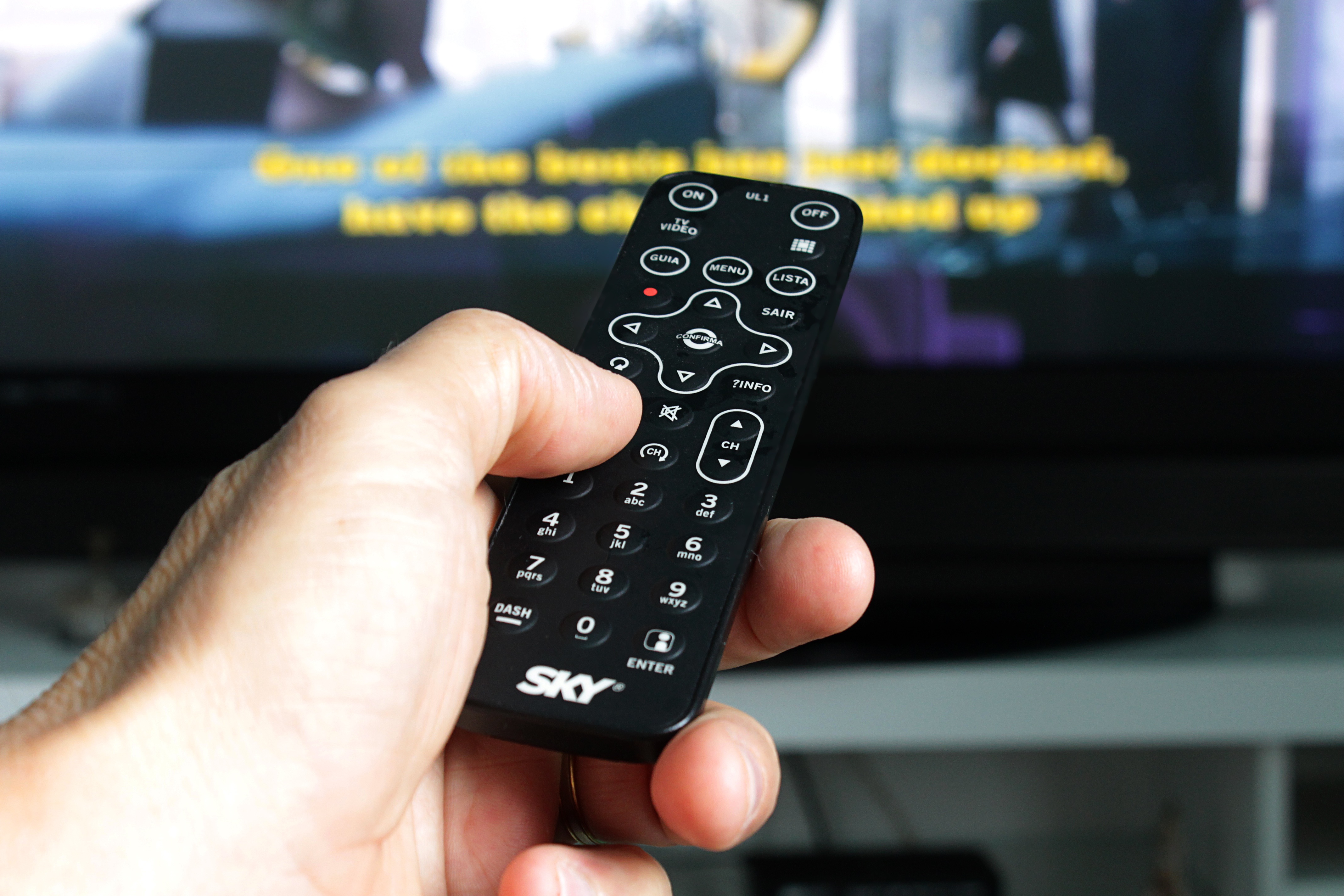 En hand håller en fjärrkontroll och en tv syns i bakgrunden.
